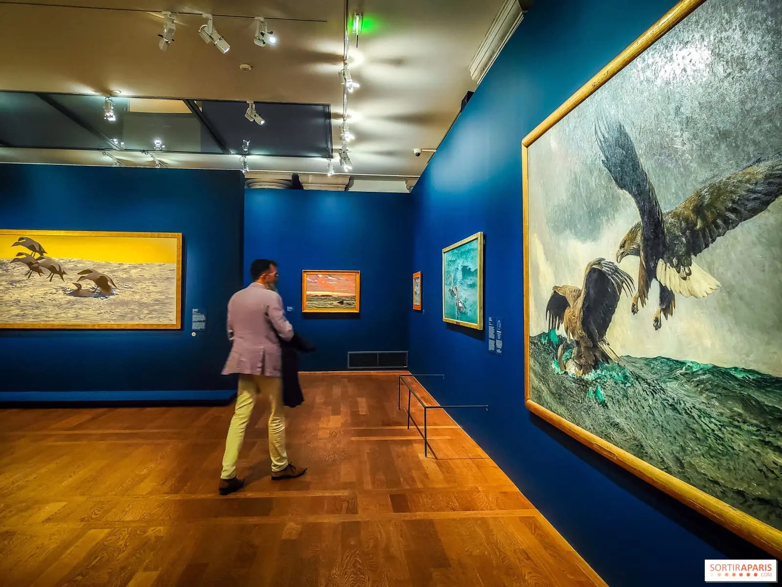 Salle de musée avec des tableaux accrochés aux murs et un visiteur qui les observe.